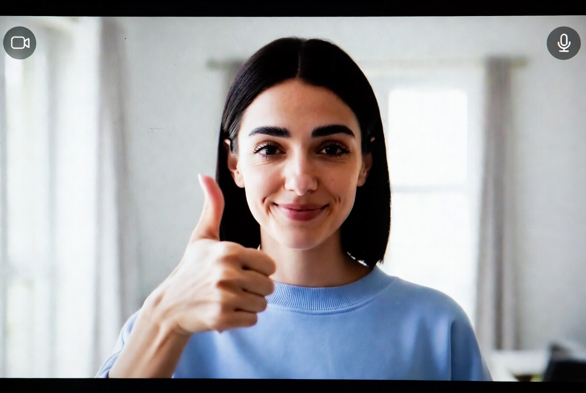 Attractive young woman giving thumbs up during live video call, wearing light blue top